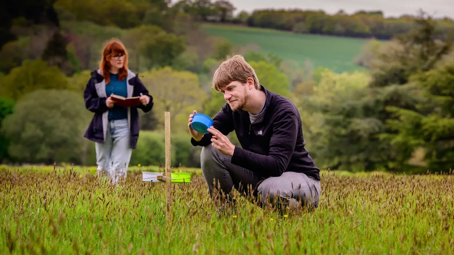 Two people working in a field, collecting seeds. One person stands with a clipboard while another examines seeds in a blue pot.