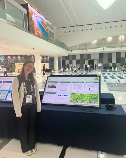A person standing beside a large digital poster display in a modern conference hall, showing research slides about plant conservation.