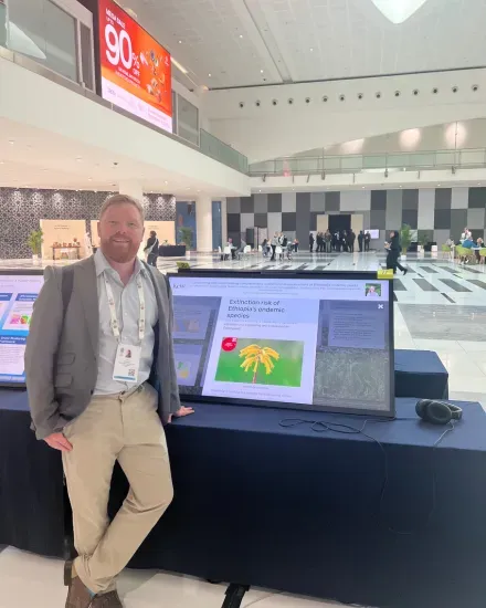 A person standing next to a large digital screen displaying plant conservation information in a bright conference venue.