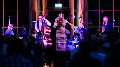 A jazz band with a singer lit in purple light in a glass conservatory performing in front of a large shadowy crowd
