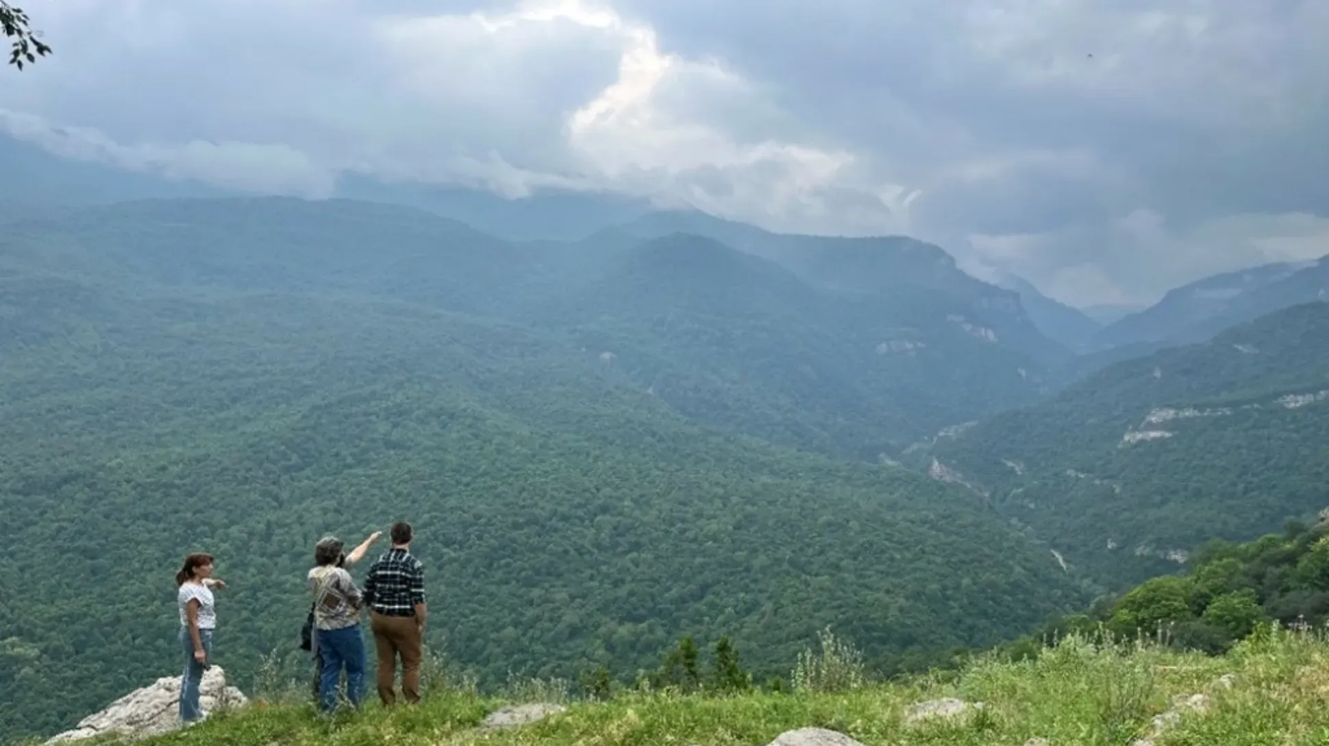 Three people overlooking a mountainous landscape covered in trees under a cloudy sky