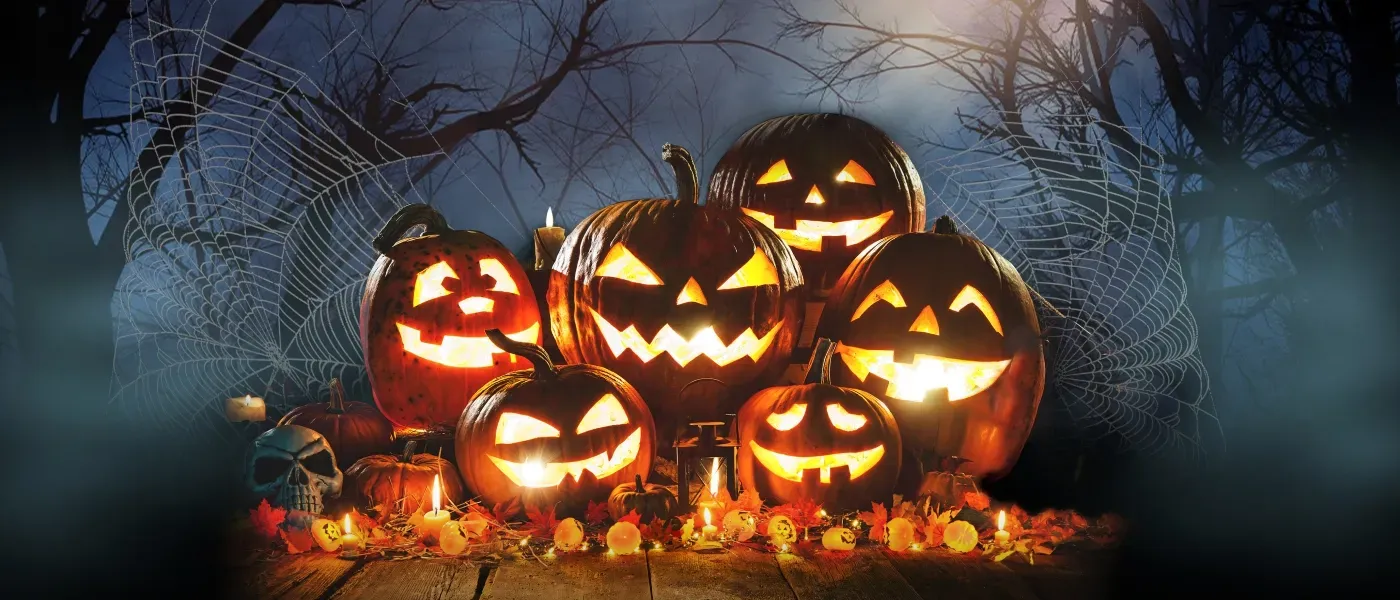 Glowing jack-o’-lanterns with carved faces arranged together on the ground, surrounded by candles, autumn leaves, and a skull decoration, with a spooky night sky, bare trees, spider webs, a full moon, and a flying bat in the background.