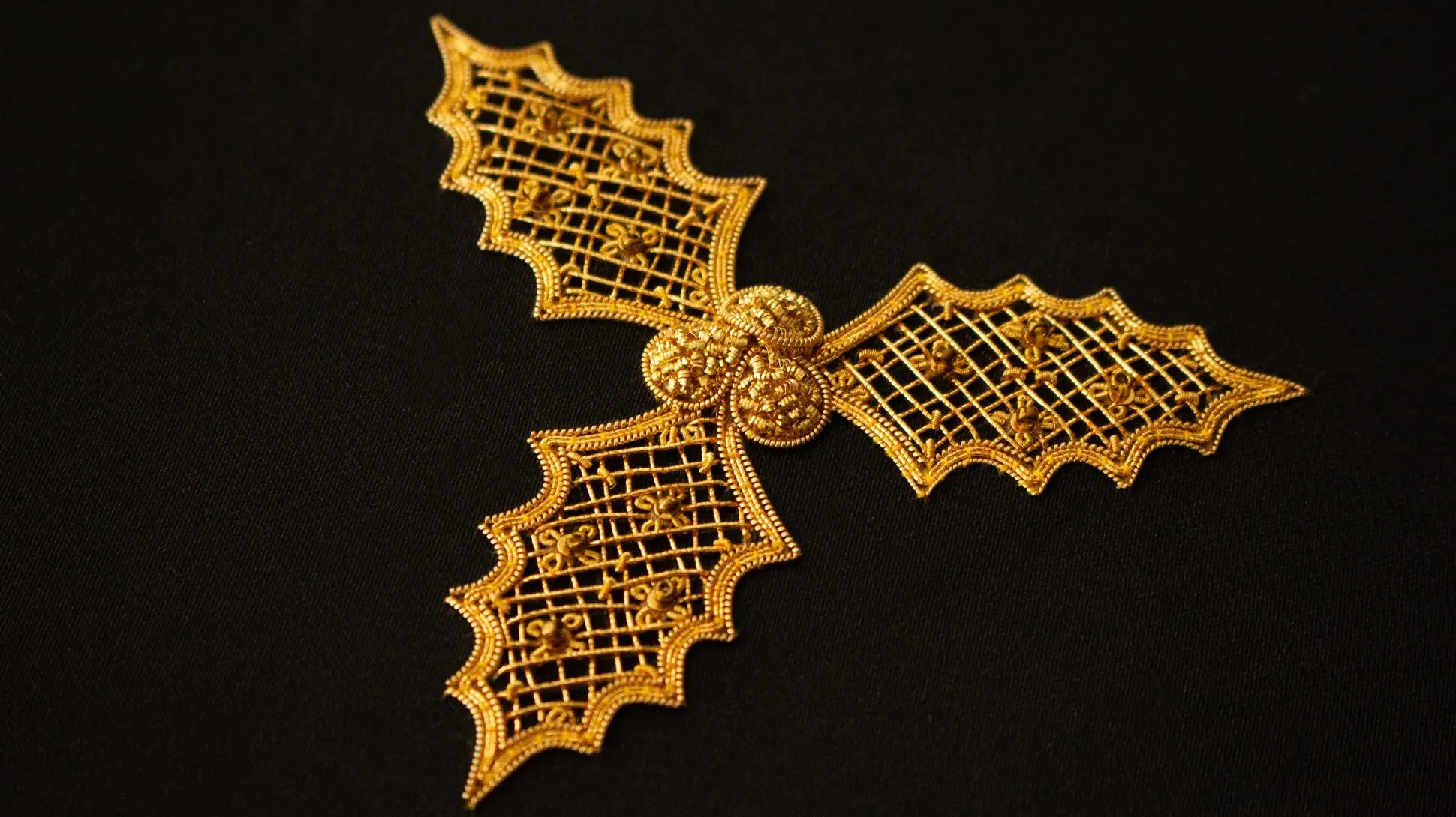 Intricate goldwork embroidery on black fabric, depicting three holly leaves with scalloped edges, joined at the center by three raised gold berries.