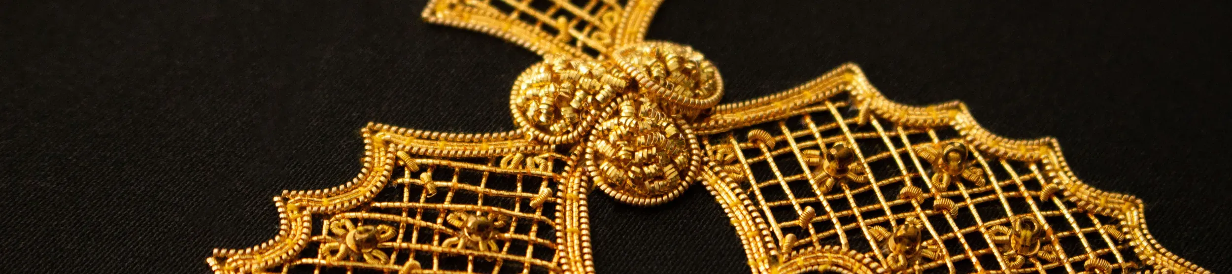 Intricate goldwork embroidery on black fabric, depicting three holly leaves with scalloped edges, joined at the center by three raised gold berries.