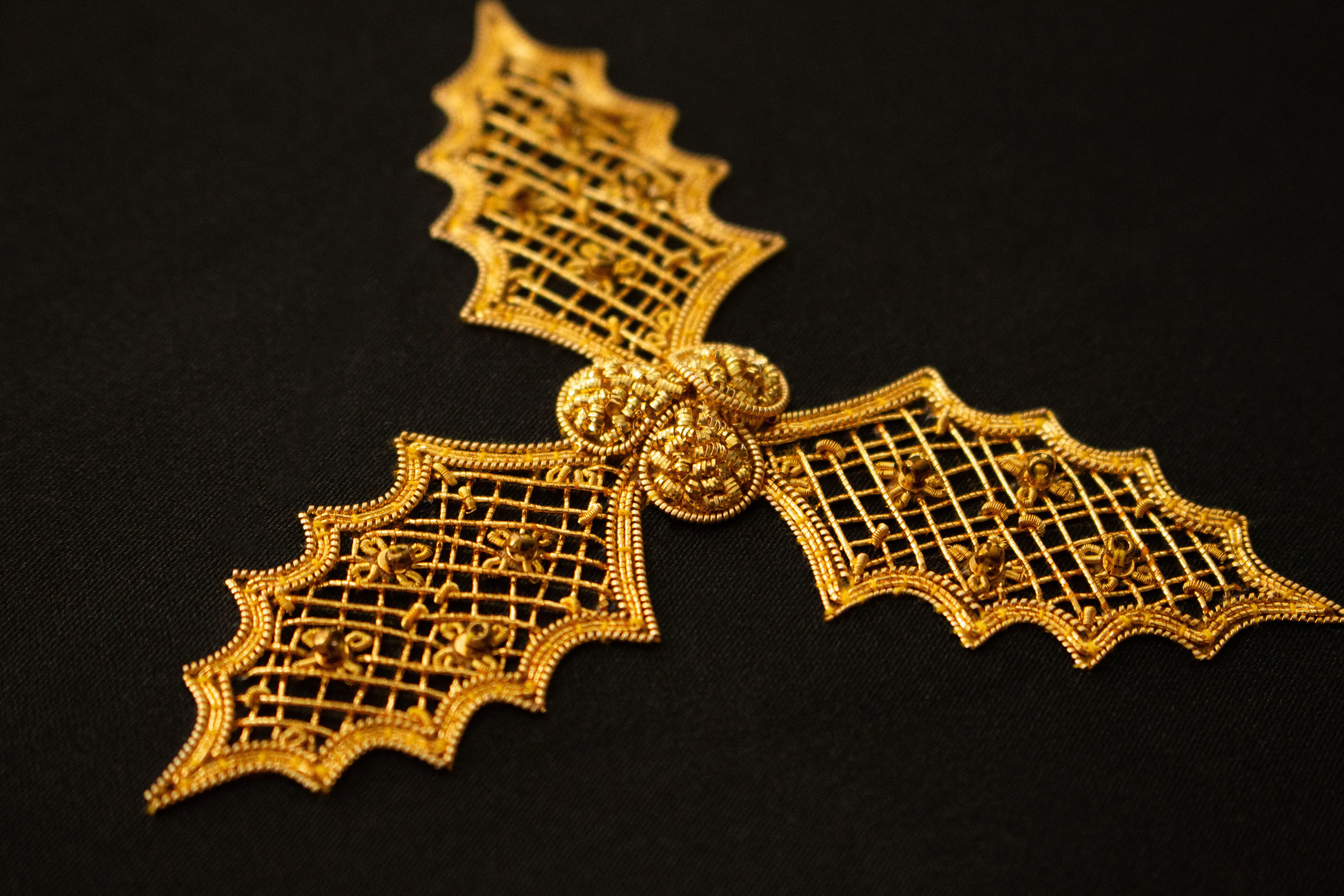 Intricate goldwork embroidery on black fabric, depicting three holly leaves with scalloped edges, joined at the center by three raised gold berries.