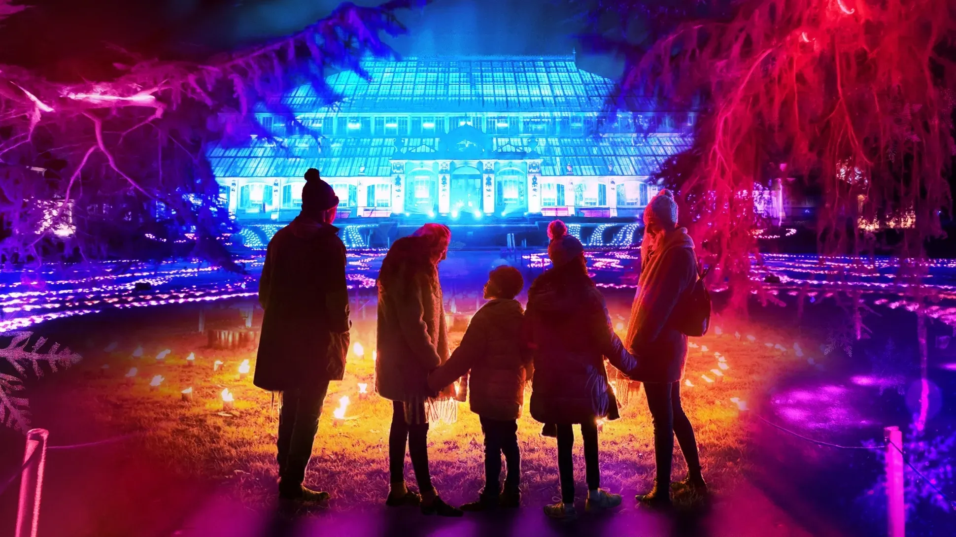 A group of two adults and three children wearing winter clothing stand in front of an illuminated Temperate House at Kew Gardens for Christmas at Kew