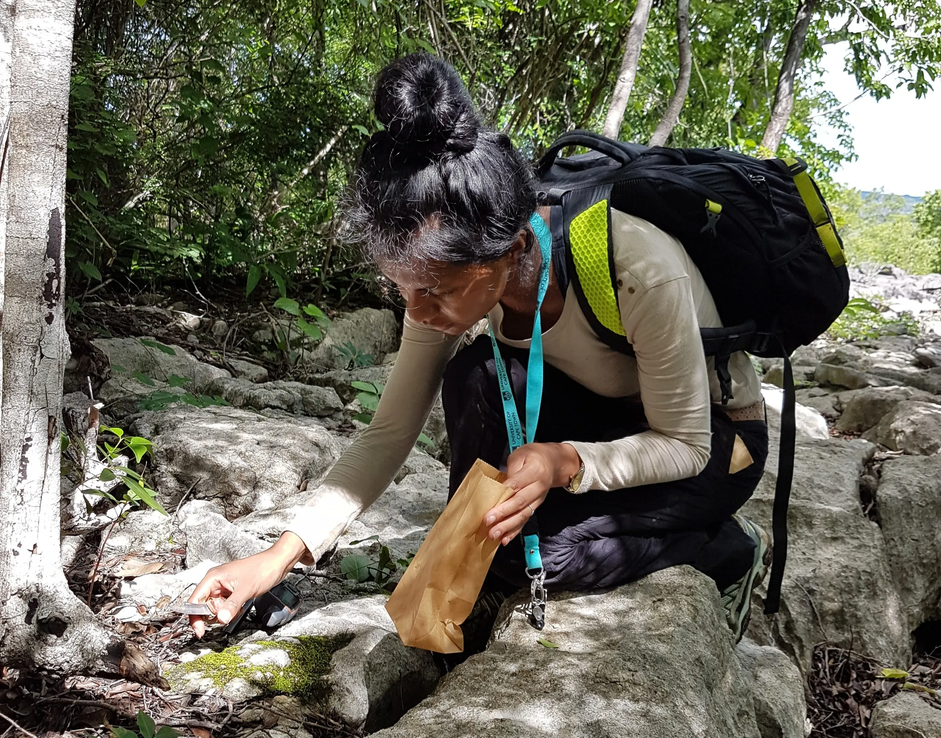 Lova crouched down among some rocks with a small knife in one hand and a paper bag in the other