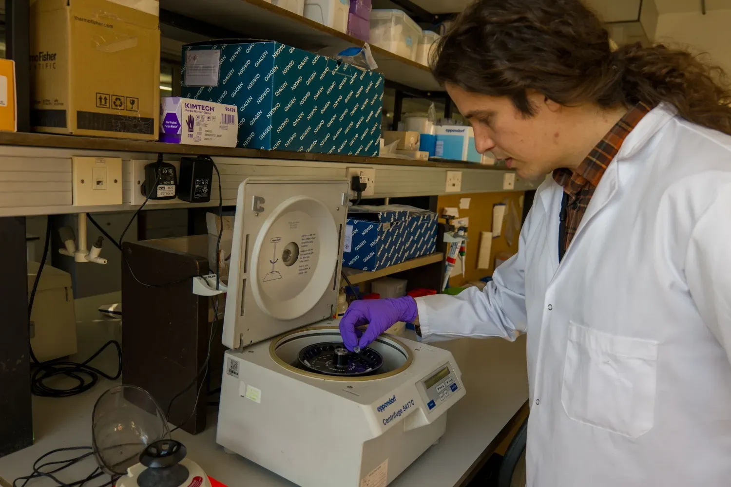A researcher in a white lab coat and purple gloves placing a sample into a centrifuge in a genomics laboratory.