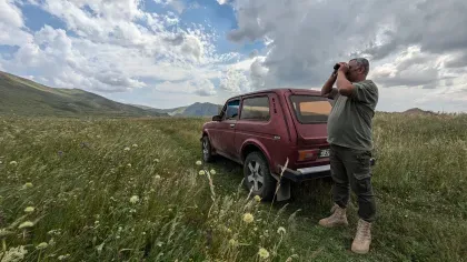 A person stood next to a car surveying the area with binoculars in a wide valley filled with meadow flowers