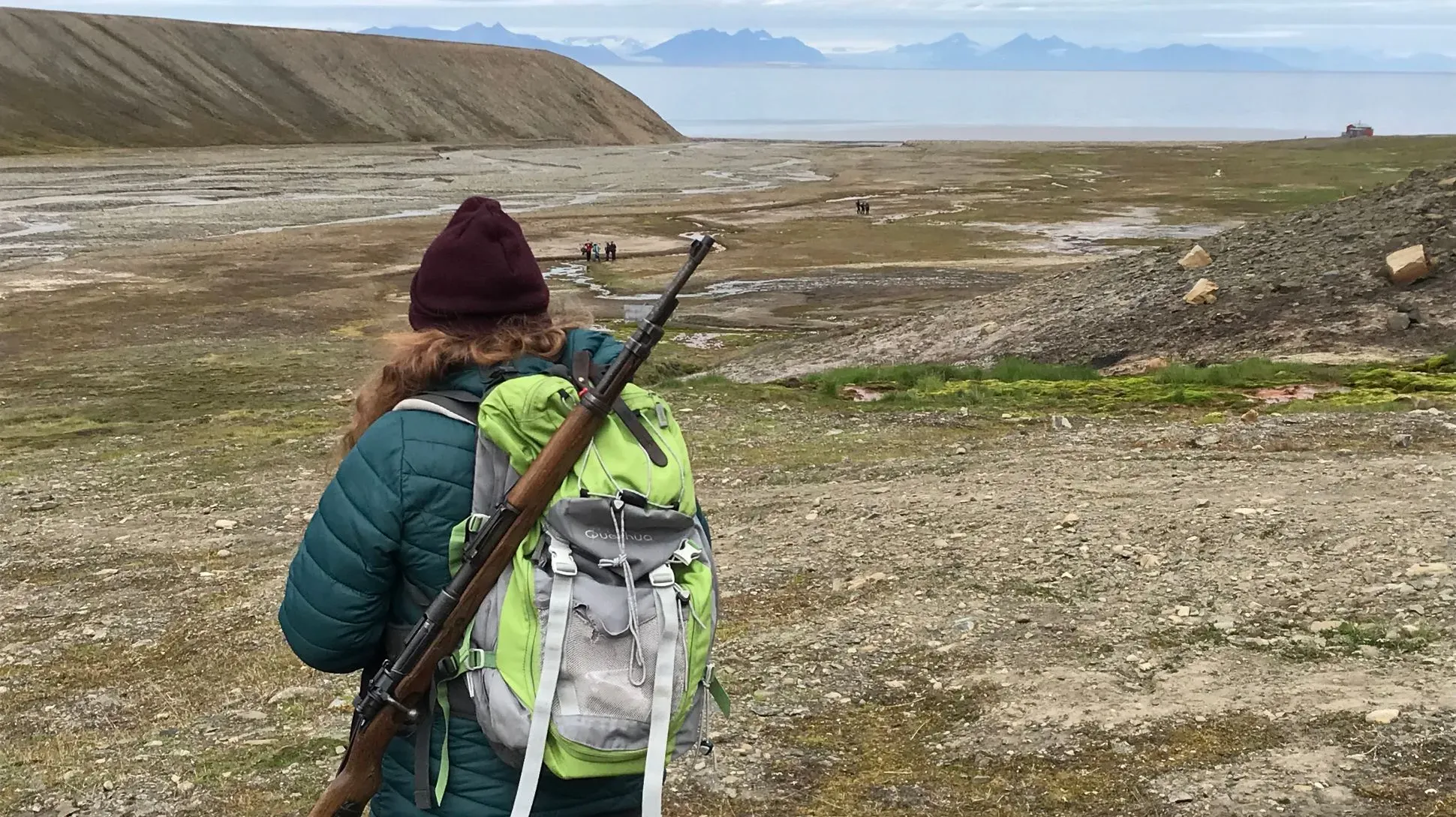 A person carrying a large backpack and a rifle walks through an open field with mountains 