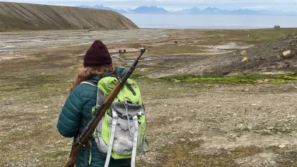 A person carrying a large backpack and a rifle walks through an open field with mountains 