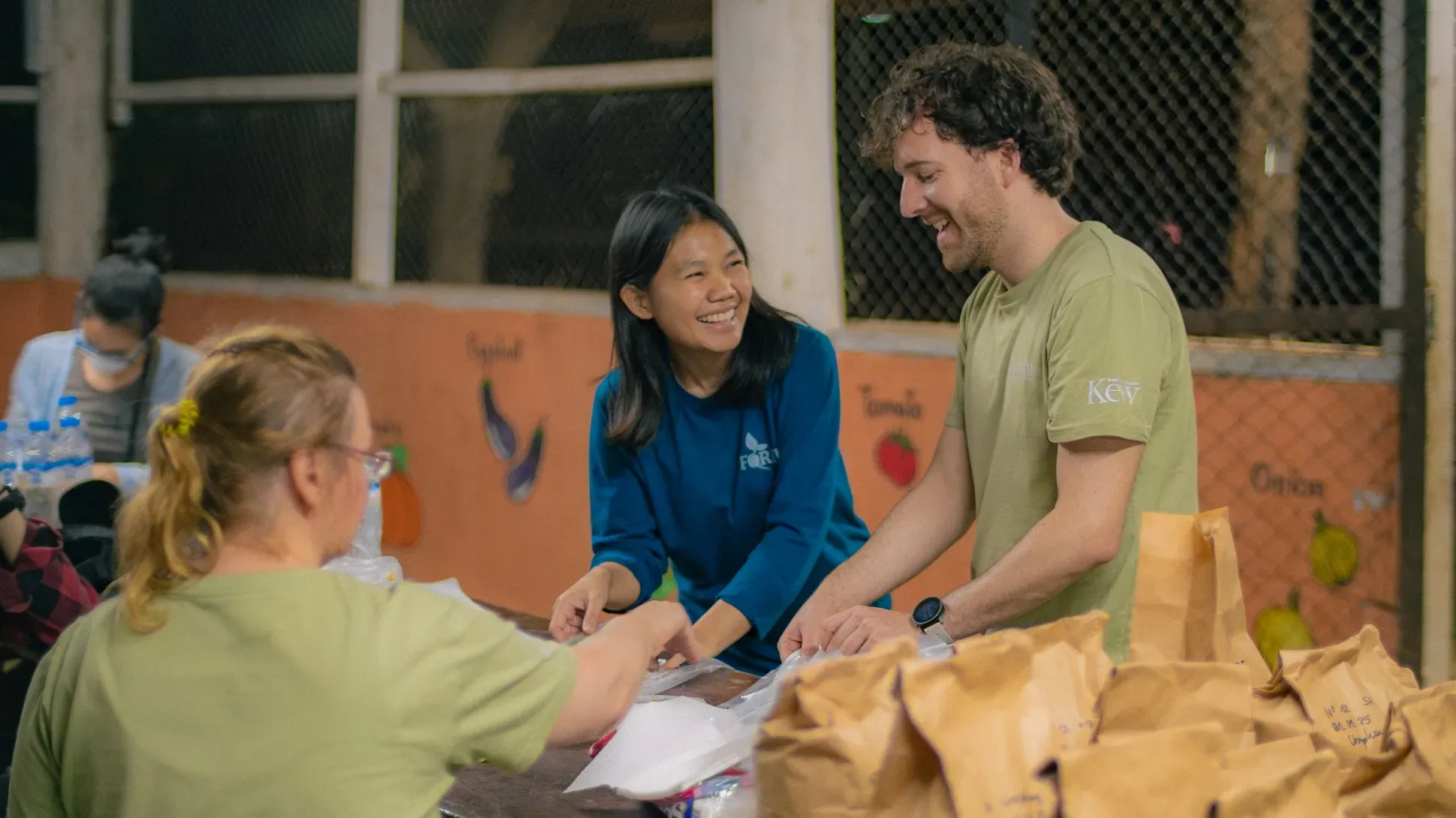 Two people smile as they sort brown paper bags on a table. There are two other people on the left hand side, one in the foreground and one in the background