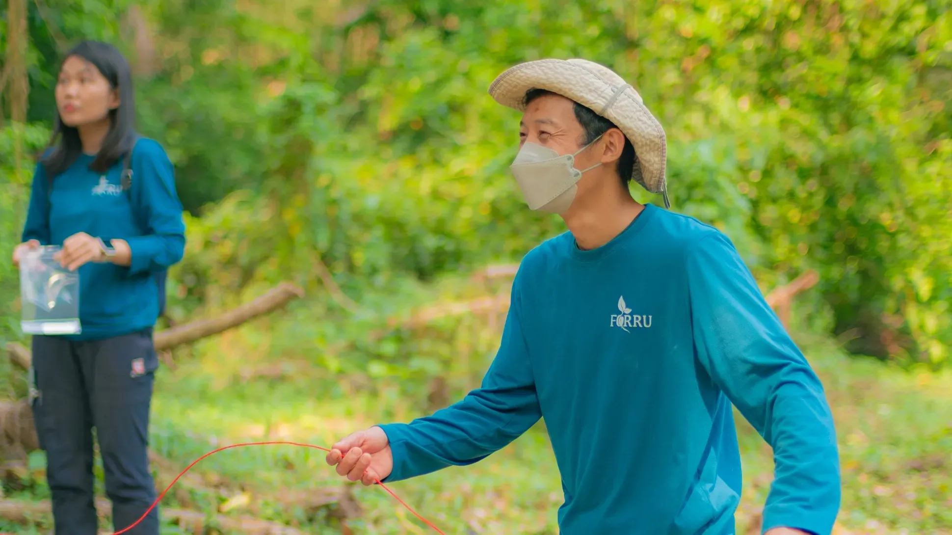 A man wearing a mask and cyan jump in a forested environment. He is throwing a rope. A person in a similar jumper stands to the back and left of him.